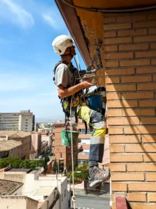 Trabajador reparando voladizo con grietas en fachada usando herramientas de albañilería